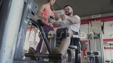人与私人教练在健身房的训练模拟器上摇动他的手臂肌肉。 个人减肥训练